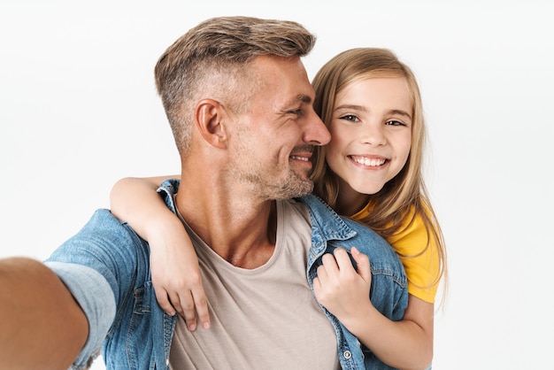 Alegre padre caucásico que lleva a cuestas a su pequeña hija mientras toma una foto selfie juntos aislado en blanco