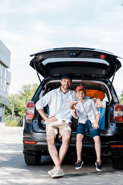 Alegre pai sentado perto do filho com biplano de madeira perto do carro