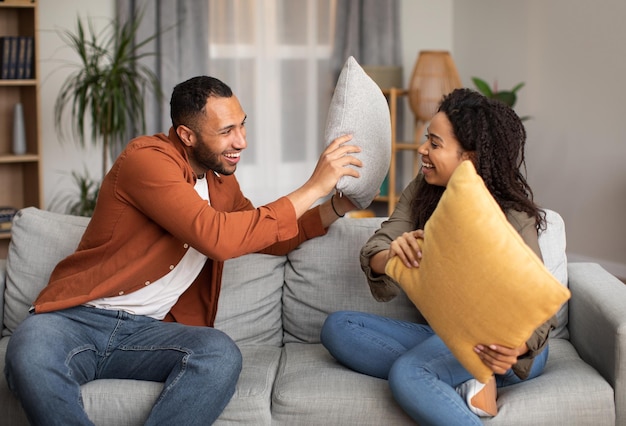 Alegre pareja africana teniendo pelea de almohadas por diversión en casa