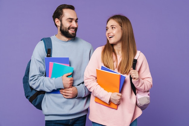 una alegre pareja amorosa amigos estudiantes aislados sobre una pared púrpura sosteniendo libros.