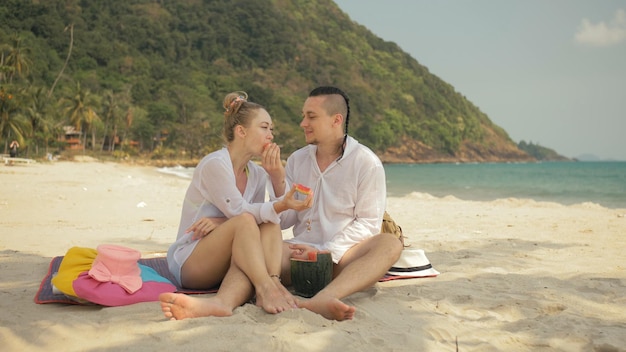 La alegre pareja amorosa sosteniendo y comiendo rebanadas de sandía en el mar tropical de la playa de arena. Amantes románticos dos personas pasan el fin de semana de verano.