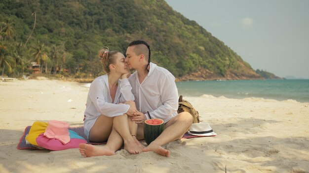 La alegre pareja amorosa sosteniendo y comiendo rebanadas de sandía en el mar tropical de la playa de arena