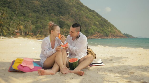 La alegre pareja amorosa sosteniendo y comiendo rebanadas de sandía en el mar tropical de la playa de arena