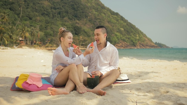 La alegre pareja amorosa sosteniendo y comiendo rebanadas de sandía en el mar tropical de la playa de arena