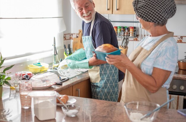 Alegre pareja de ancianos en la cocina de la casa mientras la esposa sostiene un pastel de plumcake casero recién horneado y el esposo lava los platos