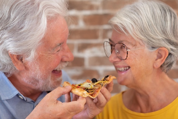 Alegre pareja de ancianos divirtiéndose juntos comiendo una pizza Anciana y hombre riéndose sentados en la mesa de casa disfrutando de una rebanada de pizza