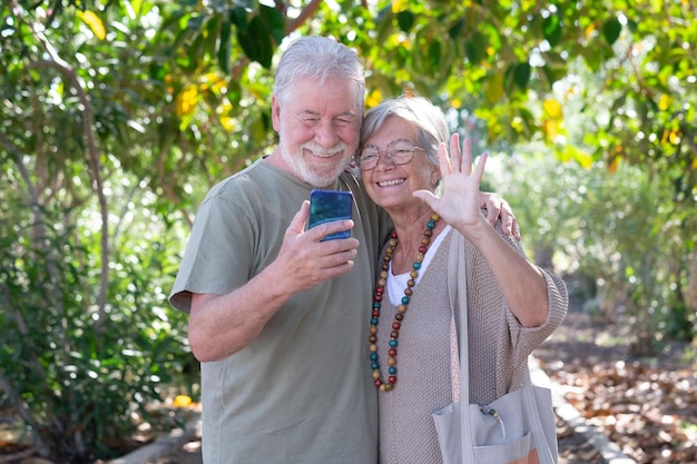 Alegre pareja de ancianos de pelo blanco caminando en el parque público mientras usa el teléfono móvil en video chat con la familia Sonriente mujer saludando con la mano mirando el teléfono celular