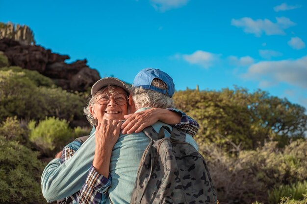 Alegre pareja caucásica mayor abrazándose en trekking de montaña disfrutando de la naturaleza y la libertad Sonrientes jubilados con sombrero y ropa informal entre arbustos verdes y cielo azul