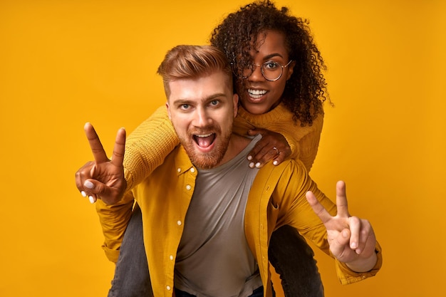 Alegre pareja diversa positiva que le muestra el signo de la victoria y da un paseo a cuestas sobre la pared naranja