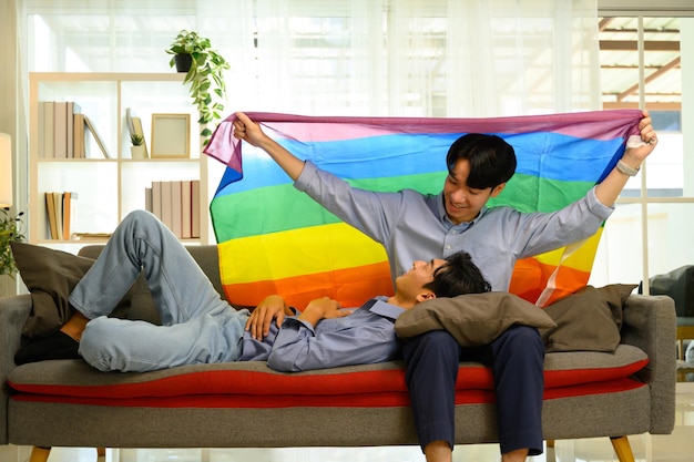 Una alegre pareja gay con una bandera del orgullo del arco iris relajándose en el sofá