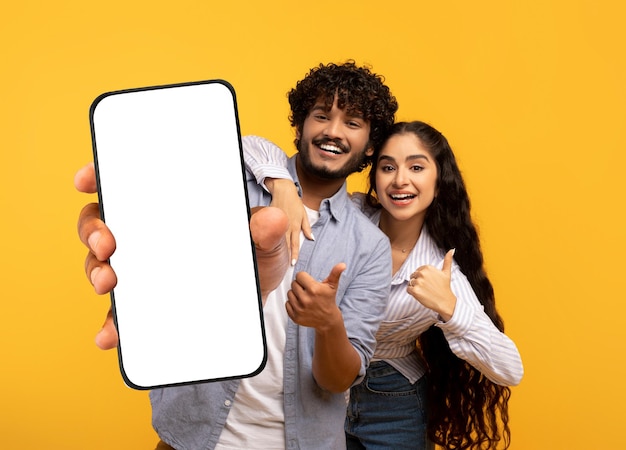 Alegre pareja india demostrando un gran teléfono inteligente en blanco y mostrando los pulgares hacia arriba