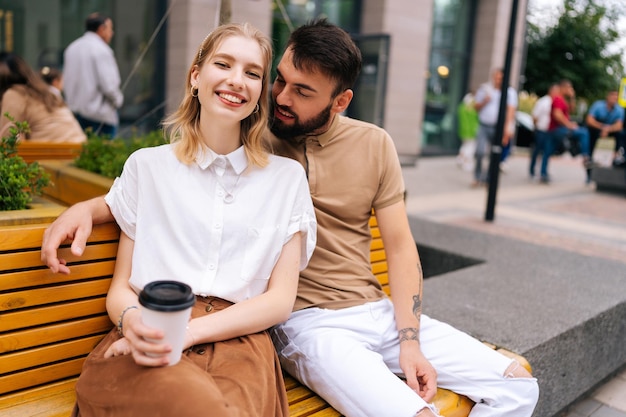 Alegre pareja joven amorosa sentada en un banco y bebiendo café para llevar disfrutando del tiempo juntos al aire libre en el día de verano