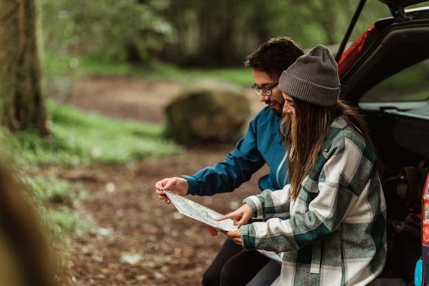 Foto una alegre pareja joven caucásica con chaquetas en el bosque disfruta del viaje sentada en el baúl mira el mapa de la ruta