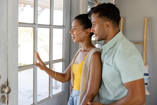 Alegre pareja joven caucásica mirando por la ventana mientras está de pie en el hotel, copie el espacio. Concepto inalterado, amor, juntos, vacaciones, centro turístico, estilo de vida y disfrute.