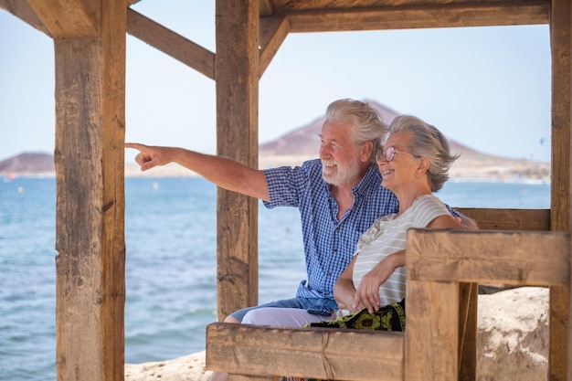 Alegre pareja mayor caucásica sentada a la sombra de la glorieta frente al mar mirando a los ancianos jubilados disfrutando de las vacaciones en el mar en un día ventoso y soleado