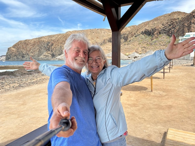 Una alegre pareja de mayores sonrientes abrazándose al aire libre juntos en la playa toman fotos con un poste de selfie