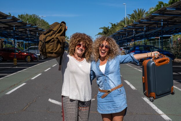 Foto alegre pareja de mujeres rizadas en el estacionamiento del aeropuerto con equipaje y mochila lista para viajar. sonriendo mirando a la cámara