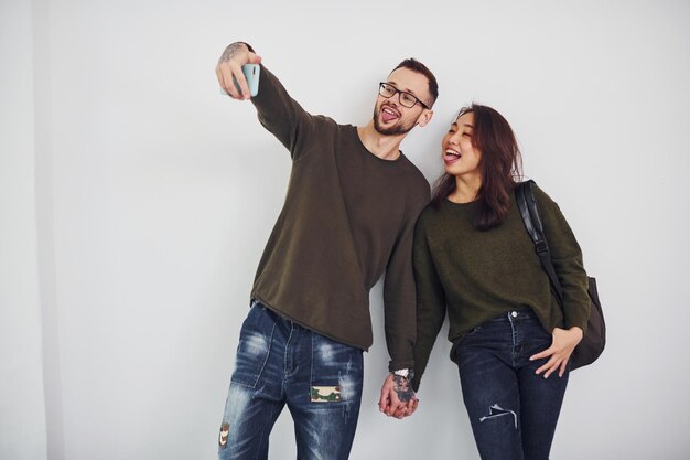 Alegre pareja multiétnica con ropa informal haciendo selfie en el interior del estudio con fondo blanco.