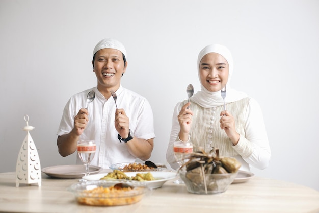 Una alegre pareja musulmana sostiene cubiertos frente a una mesa llena de platos especiales durante el Eid Mubarak