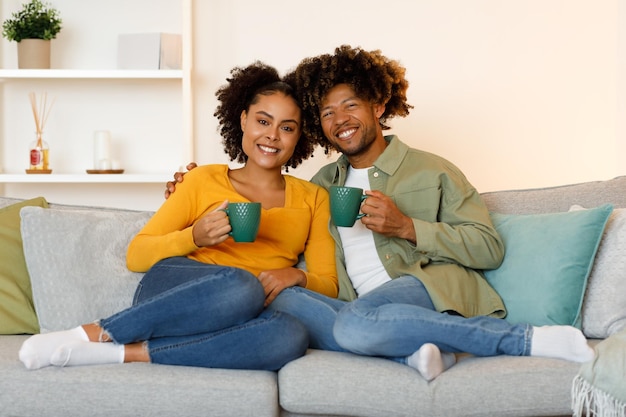Alegre pareja negra abrazándose sosteniendo tazas de café sentadas en casa