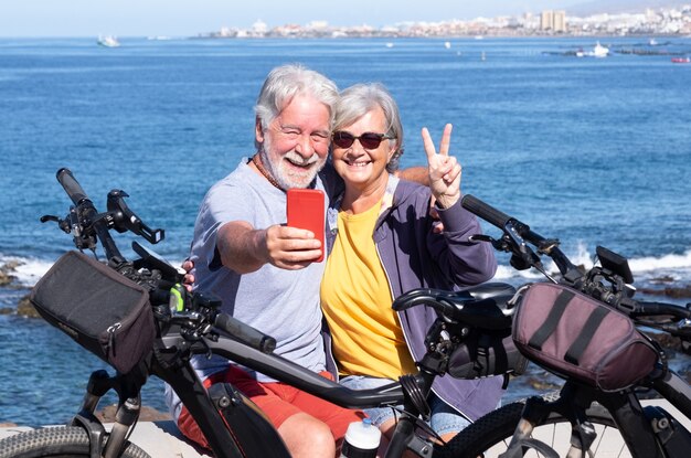 Una alegre pareja senior disfrutando de una excursión por el mar con sus bicicletas, horizonte sobre el mar. Jubilados hermosos activos que disfrutan de un estilo de vida saludable usando el teléfono móvil para un selfie