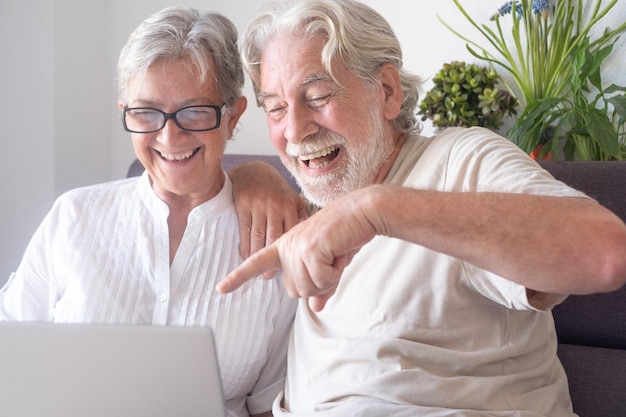 Alegre pareja senior de pelo blanco riéndose en video chat con portátil Dos jubilados caucásicos sentados en un sofá en casa disfrutando de la tecnología y las redes sociales