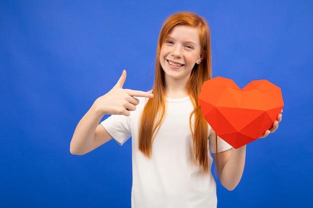 Alegre pelirroja adolescente sosteniendo un corazón rojo de papel azul con espacio de copia