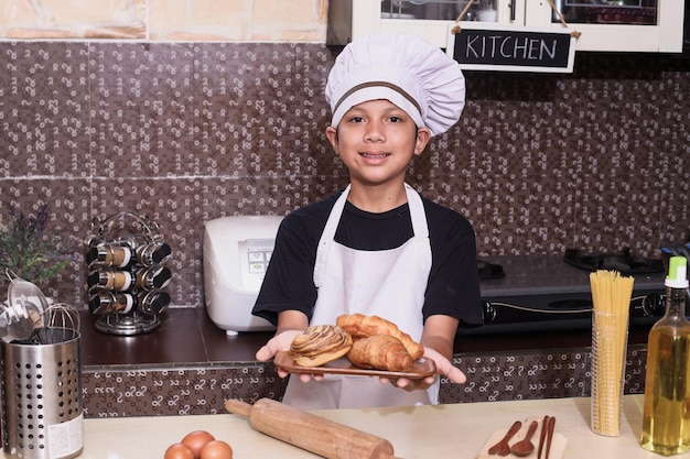 Alegre pequeno chef mostrando pão assado na placa de madeira como resultado da prática culinária na cozinha