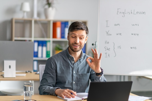 Alegre profesor masculino dando una lección de inglés en línea en una computadora portátil mostrando un gesto correcto en la oficina