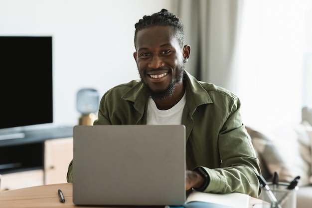 Alegre programador afroamericano sentado en el escritorio en casa freelance masculino sentado frente a la computadora