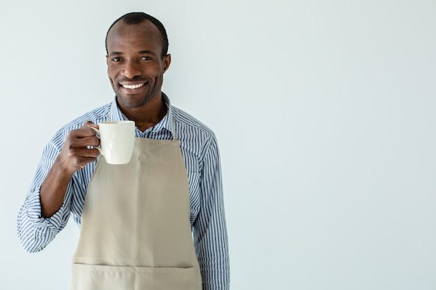 Alegre propietario de café afroamericano tomando café mientras está de pie contra la pared blanca