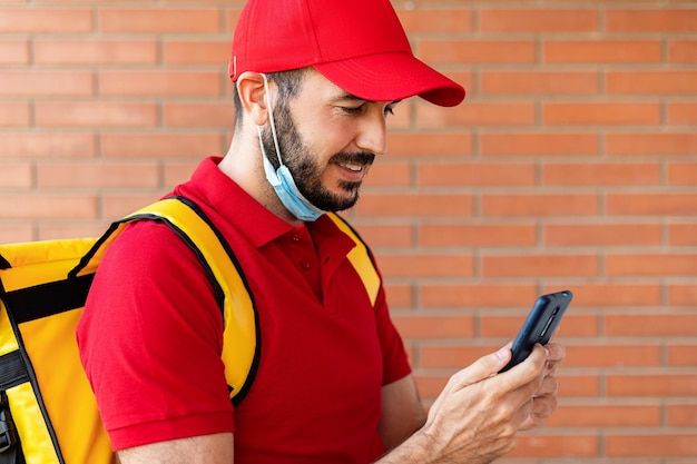 Alegre repartidor hispano con mascarilla usando un teléfono móvil en la calle mientras entrega paquetes Concepto de servicio de entrega y pequeñas empresas