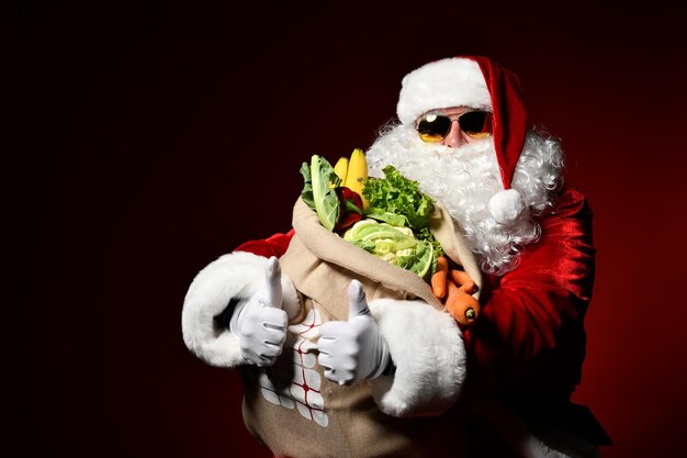 Alegre Santa Claus en gafas de sol sostiene una gran bolsa llena de frutas y verduras pulgares arriba