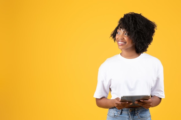 Foto alegre senhora afro-americana encaracolada milenar em camiseta branca digitando no tablet, olhe para o espaço vazio