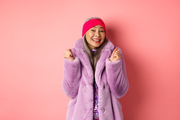 Alegre senhora asiática com um casaco elegante e descolado, comemorando a vitória ou o sucesso, dizendo sim e sorrindo feliz, em pé sobre um fundo rosa