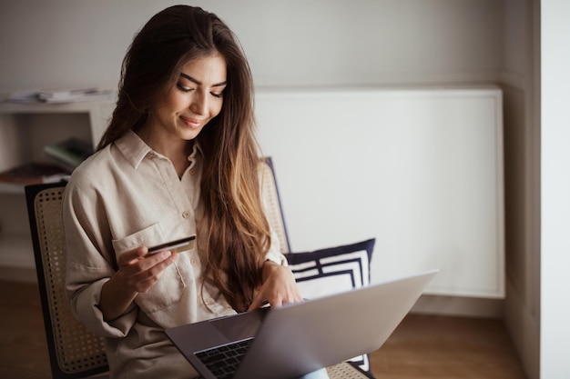 Alegre senhora de raça mista milenar digitando no laptop usando cartão de crédito, aproveite as compras on-line no quarto