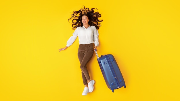 Alegre senhora posando com mala de viagem azul sobre fundo amarelo