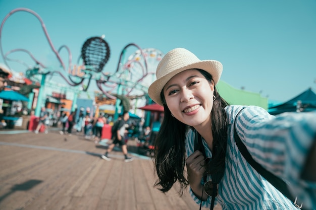 Alegre sonriente chica asiática coreana con sombrero de paja haciendo cámara de cara selfie. Retrato al aire libre de una linda mujer con estilo divirtiéndose en el parque de diversiones. mujer joven y encantadora que se toma una foto bajo el cielo azul