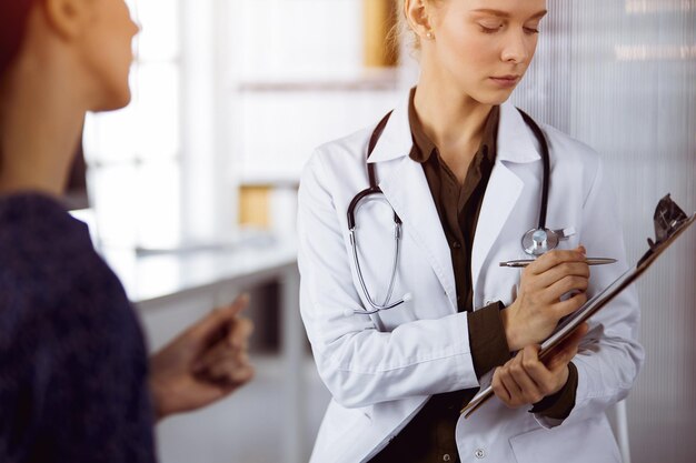 Alegre sonriente doctora y mujer paciente discutiendo el examen de salud actual mientras está sentada en una clínica soleada. Concepto de medicina.