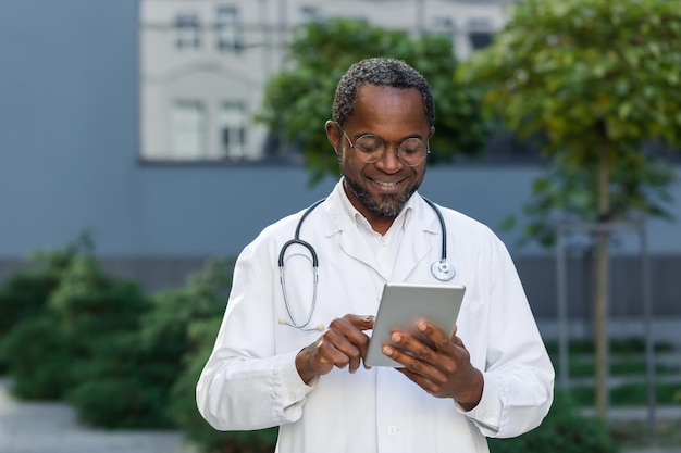 Alegre y sonriente médico senior fuera de la clínica usando una tableta hombre afroamericano usando