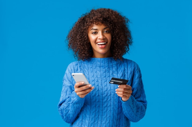 Alegre y sonriente mujer afroamericana sonriente comprando en línea, comprando durante la temporada de vacaciones con descuentos, sonriendo, sosteniendo el teléfono inteligente y la tarjeta de crédito, de pie azul