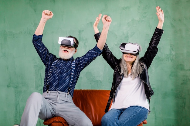 alegre sonriente pareja senior en ropa de moda elegante, sentado en una silla suave roja y disfrutando de videojuegos o películas en 3D con gafas de realidad virtual, levantando los brazos