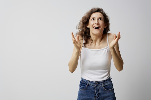 Alegre sorprendida emocionada hermosa mujer rizada en camiseta blanca básica levanta las manos ganó un millón posando aislado sobre fondo blanco Personas Emociones Concepto de estilo de vida Espacio de copia