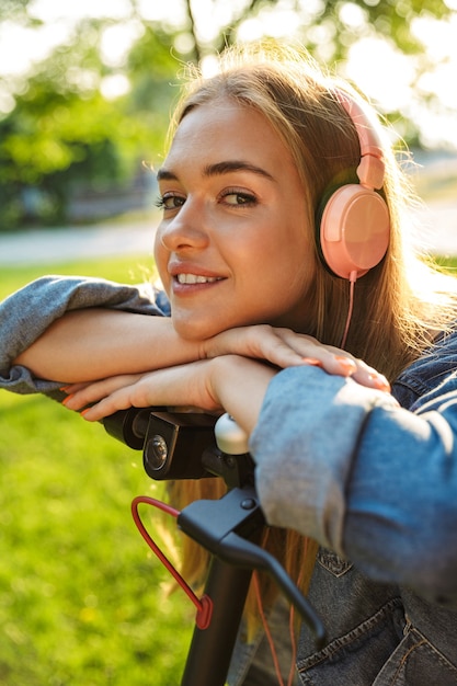 Alegre sorridente adolescente lá fora no parque verde natural na grama, ouvindo música com fones de ouvido com scooter.