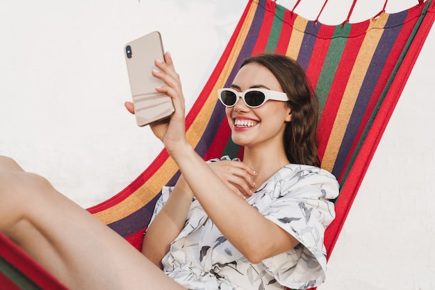 alegre sorridente jovem bela mulher na praia, posando em uma rede, tirar uma selfie pelo celular.