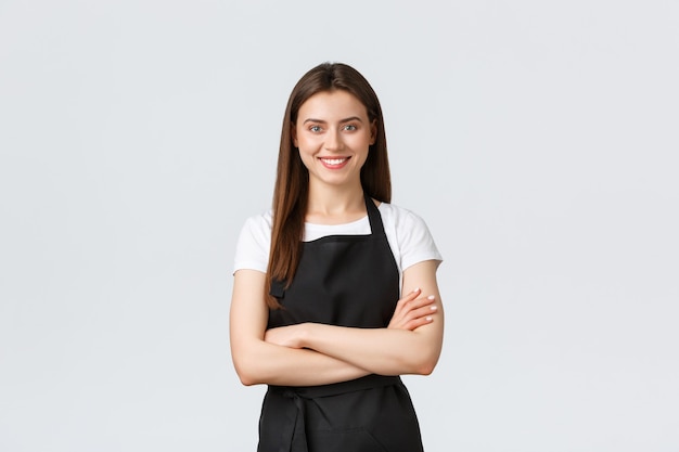 Alegre sorridente mulher barista em avental preto no peito de braços cruzados, parecendo pronta e confiante. Empregada jovem abre a cafeteria, cumprimentando os clientes. Loja de execução de vendedora.