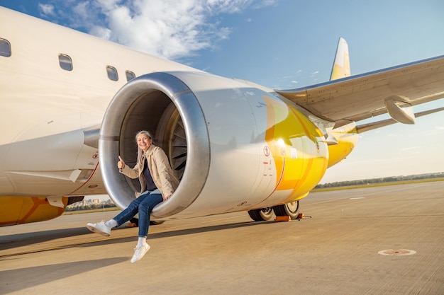 Alegre viajante feminina mostrando gesto de aprovação e sorrindo enquanto descansava no motor de turbina de avião no aeroporto