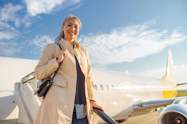 Alegre viajera mirando a la cámara y sonriendo mientras está de pie en las escaleras de embarque de aviones bajo un cielo nublado