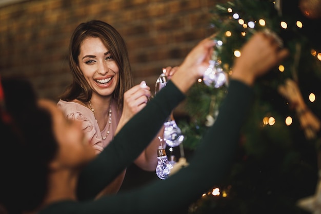 Alegres amigas multiétnicas divirtiéndose y decorando el árbol de Navidad mientras pasan las vacaciones en casa.