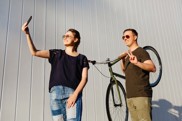 Foto alegres amigos se divertindo juntos. uma linda mulher toma uma selfie no smartphone, em pé com cara feliz, segurando uma bicicleta no ombro e mostrando sinal de paz. ao ar livre.